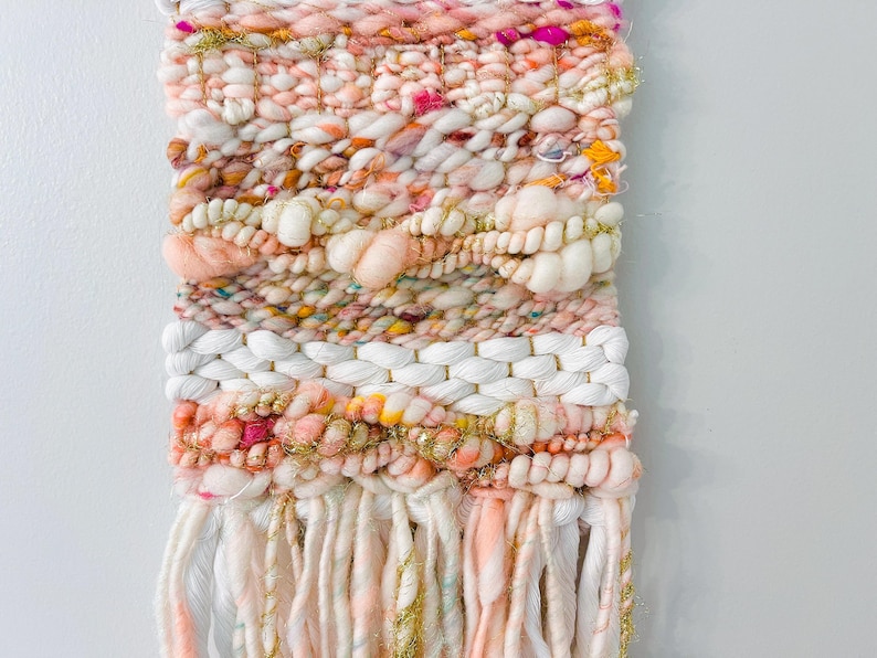 May include: A pink, white, and gold woven wall hanging with fringe. The wall hanging is made with a variety of textures and colors, including fluffy yarn, sparkly thread, and woven strands.
