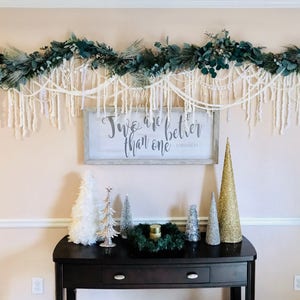 May include: A white sign with the text "Two are better than one" hangs on a wall decorated with a green garland and white yarn. A black console table with a drawer sits in front of the wall, decorated with small artificial Christmas trees.