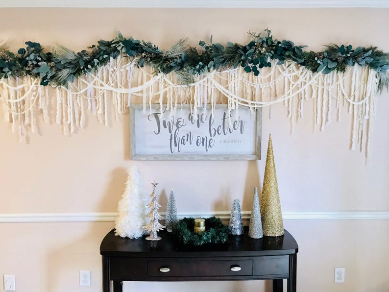 May include: A white sign with the text "Two are better than one" hangs on a wall decorated with a green garland and white yarn. A black console table with a drawer sits in front of the wall, decorated with small artificial Christmas trees.