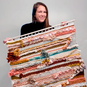 May include: A large woven wall hanging with a variety of textures and colors, including pink, white, brown, and turquoise. The hanging is made with yarn and has a wooden dowel at the top.