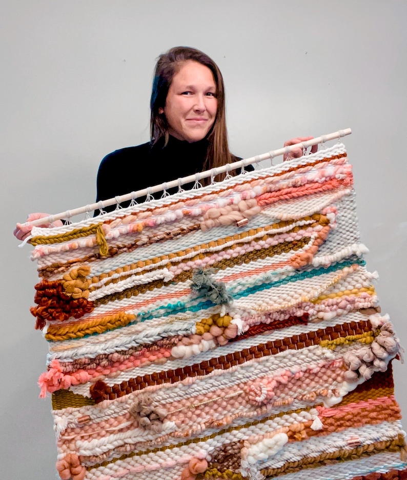 May include: A large woven wall hanging with a variety of textures and colors, including pink, white, brown, and turquoise. The hanging is made with yarn and has a wooden dowel at the top.