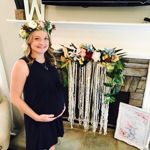 May include: A pregnant woman wearing a dark blue dress and a floral crown stands in front of a fireplace decorated with a floral garland and a white yarn macrame hanging. A framed sign with the text "Welcome Baby Little" and a pink and gray elephant illustration is on the floor.