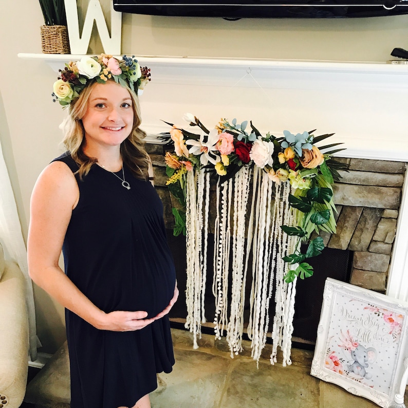 May include: A pregnant woman wearing a dark blue dress and a floral crown stands in front of a fireplace decorated with a floral garland and a white yarn macrame hanging. A framed sign with the text "Welcome Baby Little" and a pink and gray elephant illustration is on the floor.