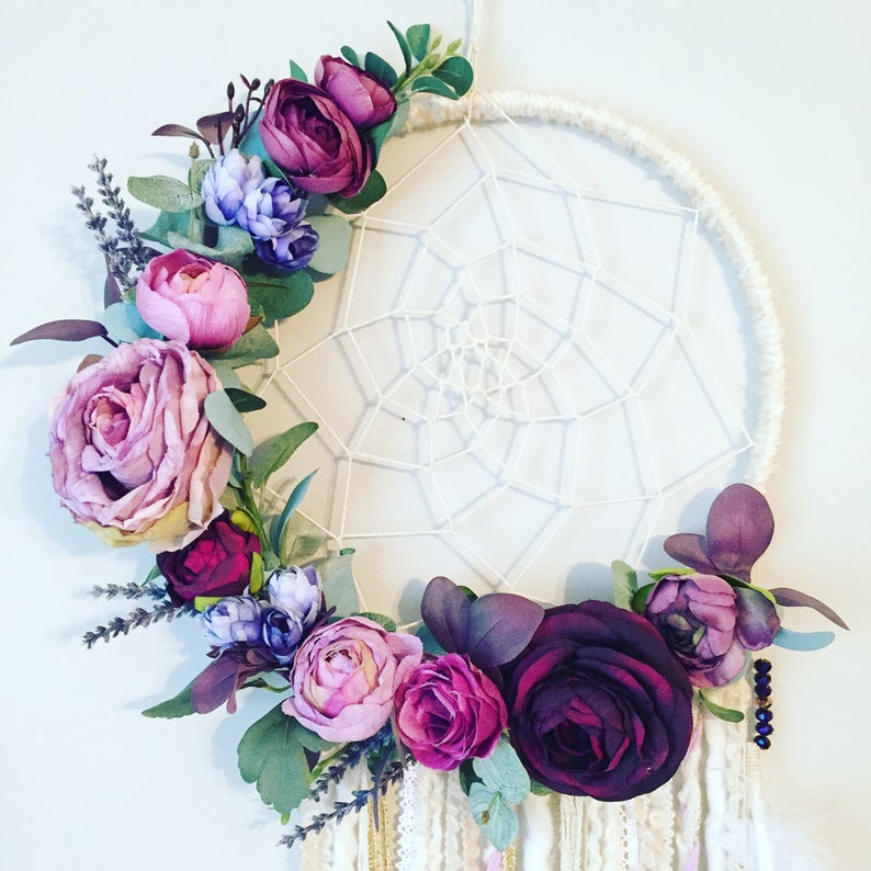 May include: A white dream catcher with a circular frame and a web-like design. The frame is decorated with a garland of purple, pink, and burgundy flowers.