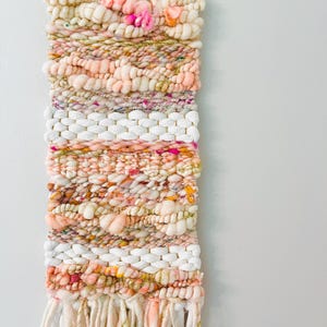 May include: A woven wall hanging with a pink, white, and gold color scheme. The hanging has a variety of textures and is made with yarn and string. It is hung on a wooden dowel rod.