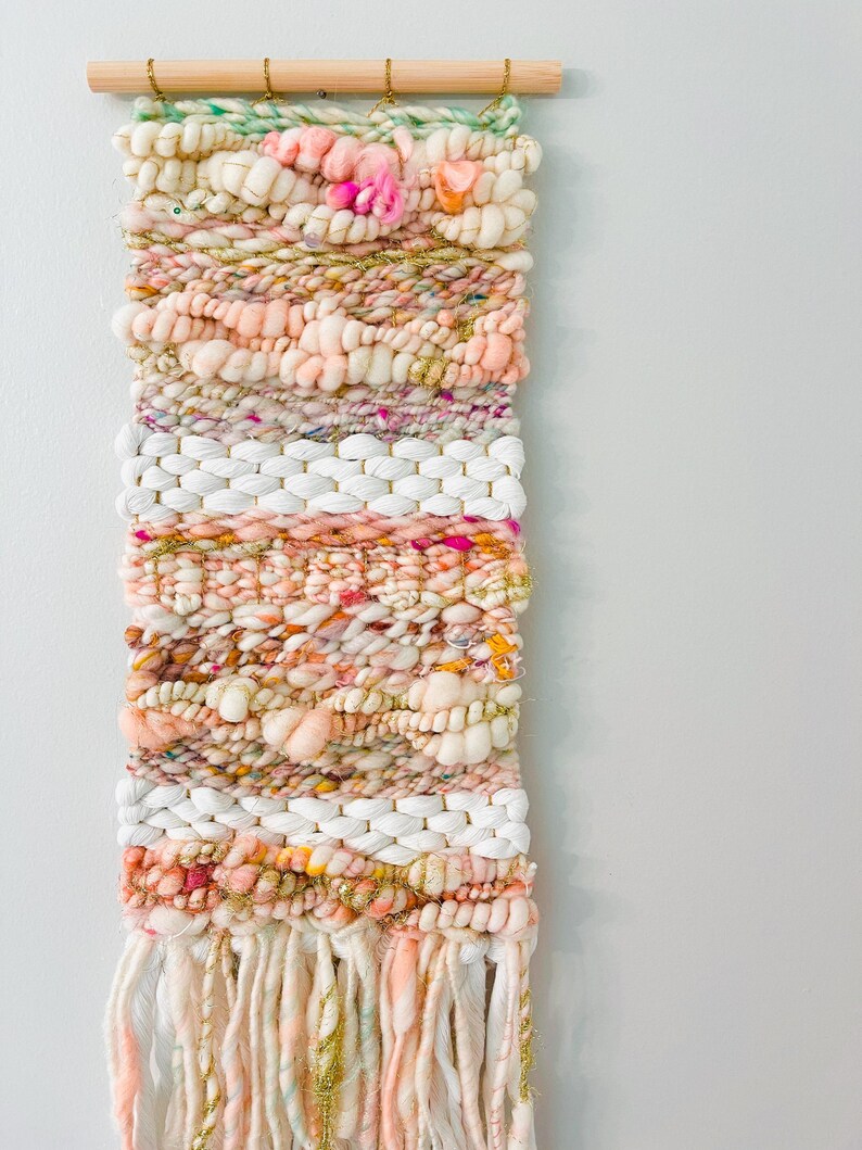 May include: A woven wall hanging with a pink, white, and gold color scheme. The hanging has a variety of textures and is made with yarn and string. It is hung on a wooden dowel rod.