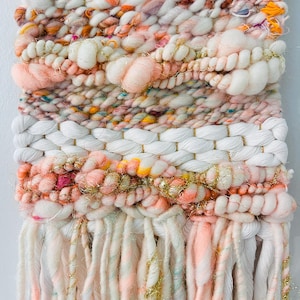 May include: A pink, white, and gold woven wall hanging with a fringe of yarn. The wall hanging has a textured, woven pattern and is decorated with gold glitter.