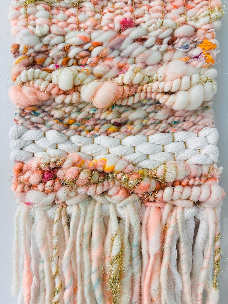 May include: A pink, white, and gold woven wall hanging with a fringe of yarn. The wall hanging has a textured, woven pattern and is decorated with gold glitter.
