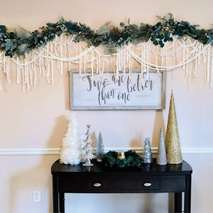 May include: A white wooden sign with the text "Two are better than one" in black calligraphy. The sign is hanging on a wall decorated with a garland of green leaves and white yarn. A black console table with a green wreath and small white and gold Christmas trees sits in front of the sign.