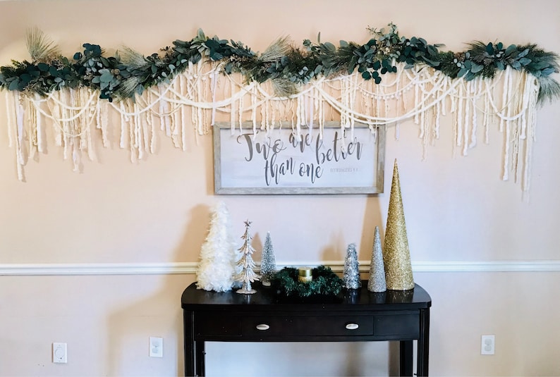 May include: A white wooden sign with the text "Two are better than one" in black calligraphy. The sign is hanging on a wall decorated with a garland of green leaves and white yarn. A black console table with a green wreath and small white and gold Christmas trees sits in front of the sign.
