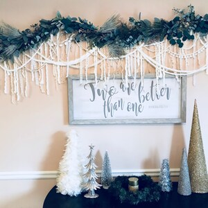 May include: A white sign with the text "Two are better than one" in grey lettering, with a garland of greenery and white fringe above it. There are several small artificial Christmas trees in front of the sign, including a white feather tree, a silver glitter tree, and a silver cone tree.