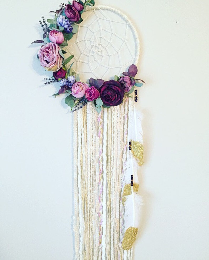 May include: A white dream catcher with a floral wreath made of purple, pink, and green flowers. The dream catcher has white and gold feathers hanging from the bottom.