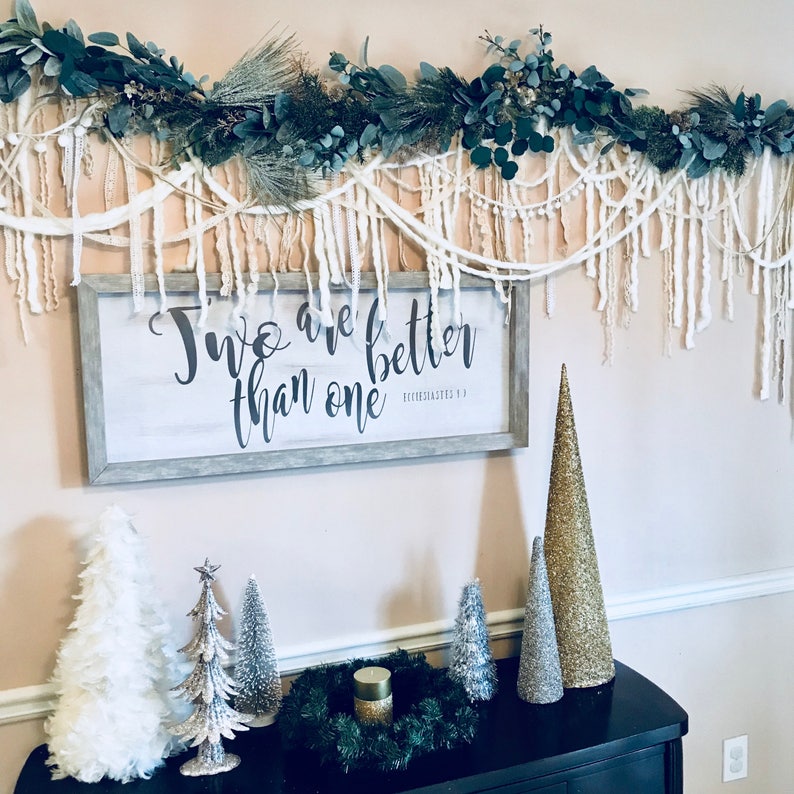 May include: A white sign with the text "Two are better than one" and a scripture reference. The sign is hanging on a wall with a white and green garland above it. There are several small Christmas trees on a black table below the sign.