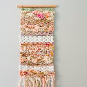 May include: A woven wall hanging with a peach, white, and pink color scheme. The hanging has a fringe at the bottom and is made with a variety of textures.
