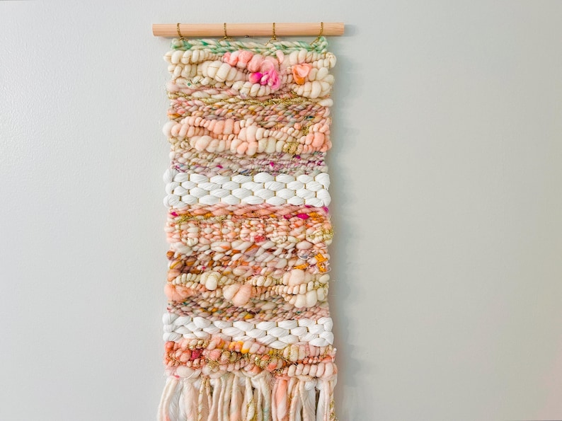 May include: A woven wall hanging with a peach, white, and pink color scheme. The hanging has a fringe at the bottom and is made with a variety of textures.