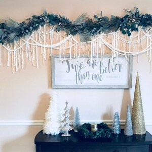 May include: A white sign with the text "Two are better than one" in grey lettering, hanging on a wall with a white garland of greenery and white yarn.  There are several small artificial Christmas trees on a black table below the sign.