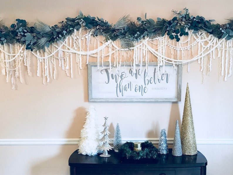 May include: A white sign with the text "Two are better than one" in grey lettering, hanging on a wall with a white garland of greenery and white yarn.  There are several small artificial Christmas trees on a black table below the sign.