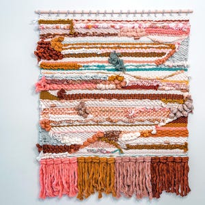 May include: A colorful woven wall hanging with a variety of textures and colors, including pink, brown, white, and blue. The hanging has a fringe border and is made with a variety of yarns.