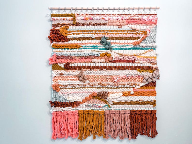 May include: A colorful woven wall hanging with a variety of textures and colors, including pink, brown, white, and blue. The hanging has a fringe border and is made with a variety of yarns.