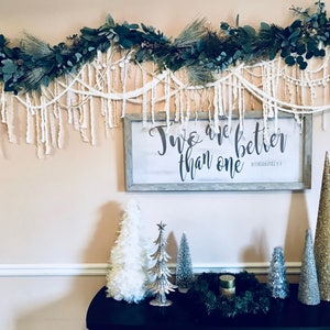 May include: A white wall with a green and white garland hanging above a wooden sign with the text "Two are better than one" in black lettering. Below the sign are five small artificial Christmas trees, three in silver, one in gold, and one in white. The trees are on a black table with a green wreath and a candle in the centre.