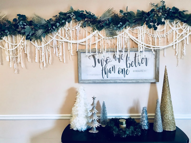 May include: A white wall with a green and white garland hanging above a wooden sign with the text "Two are better than one" in black lettering. Below the sign are five small artificial Christmas trees, three in silver, one in gold, and one in white. The trees are on a black table with a green wreath and a candle in the centre.