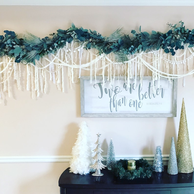 May include: A white sign with the text "Two are better than one" in grey lettering. The sign is hanging on a wall with a garland of green leaves and white yarn hanging above it. There are several small artificial Christmas trees on a dark brown table below the sign.