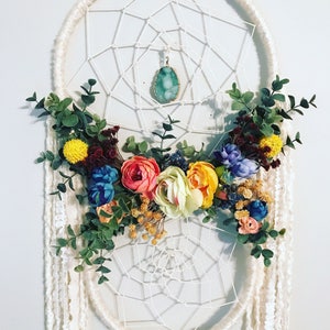Puede incluir: Un atrapasue&ntilde;os blanco con un colgante de &aacute;gata verde y una corona floral de flores artificiales de colores.