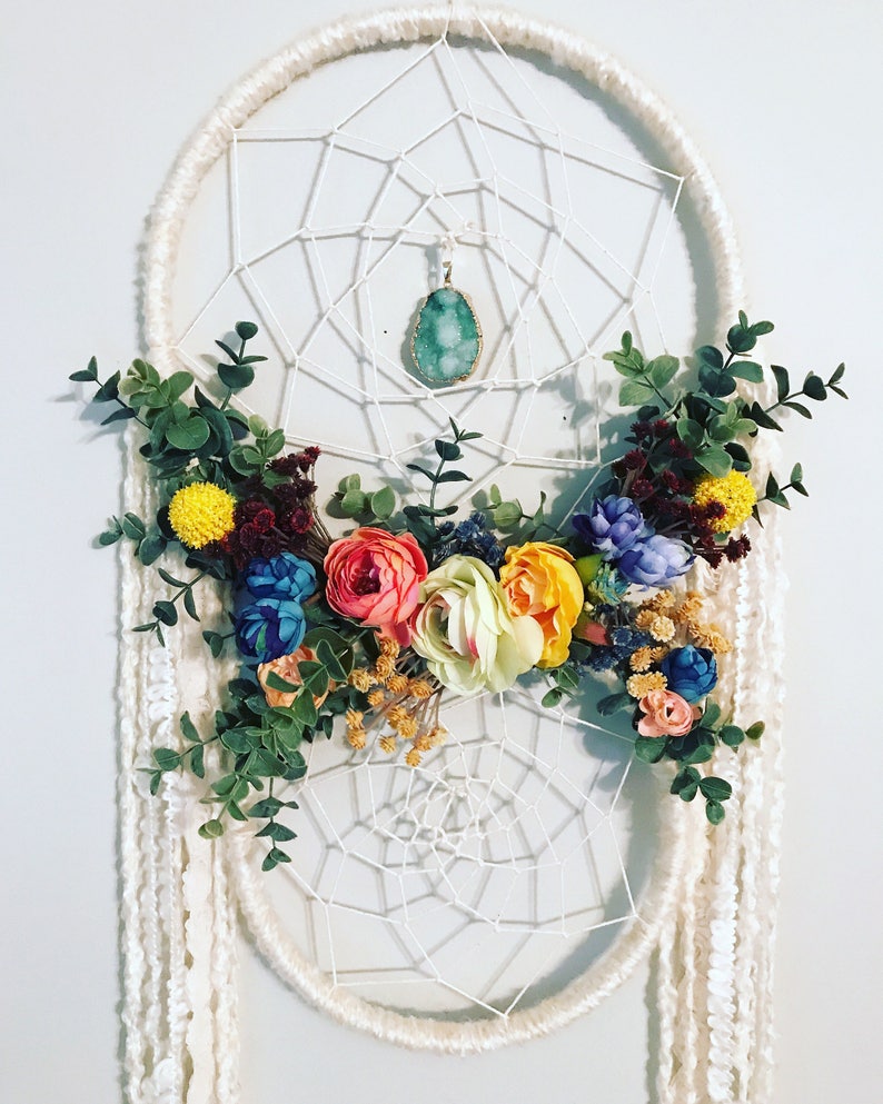 Puede incluir: Un atrapasue&ntilde;os blanco con un colgante de &aacute;gata verde y una corona floral de flores artificiales de colores.
