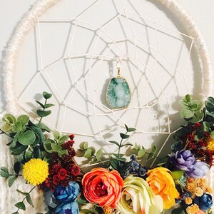 Puede incluir: Un atrapasue&ntilde;os blanco con un colgante de cristal verde y una guirnalda floral de flores amarillas, naranjas, rojas, azules y moradas.