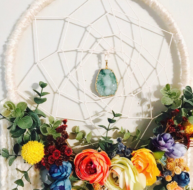 Puede incluir: Un atrapasue&ntilde;os blanco con un colgante de cristal verde y una guirnalda floral de flores amarillas, naranjas, rojas, azules y moradas.
