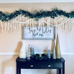 May include: A white sign with black lettering that reads "Two are better than one" with a decorative garland of greenery and white yarn hanging above it. The garland is hanging above a black console table with a variety of small, decorative trees.