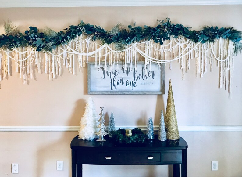 May include: A white sign with black lettering that reads "Two are better than one" with a decorative garland of greenery and white yarn hanging above it. The garland is hanging above a black console table with a variety of small, decorative trees.