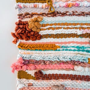 May include: A colorful woven wall hanging with a variety of textures and patterns. The hanging features a mix of pink, white, brown, and gold yarn.