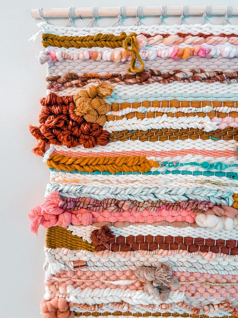 May include: A colorful woven wall hanging with a variety of textures and patterns. The hanging features a mix of pink, white, brown, and gold yarn.