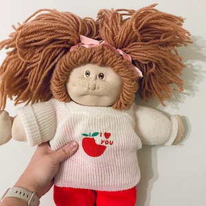May include: A vintage Cabbage Patch Kid doll with red yarn hair, wearing a white sweater with a red apple and "I love you" text, and red trousers.