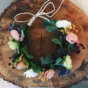 May include: A floral crown made with white, pink, and cream flowers, green leaves, and brown stems. The crown is tied with a beige string.