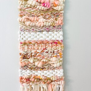 May include: A woven wall hanging with a pink, white, and gold color scheme. The hanging features a variety of textures, including fluffy yarn, chunky yarn, and thin yarn. The hanging is finished with a fringe of yarn.