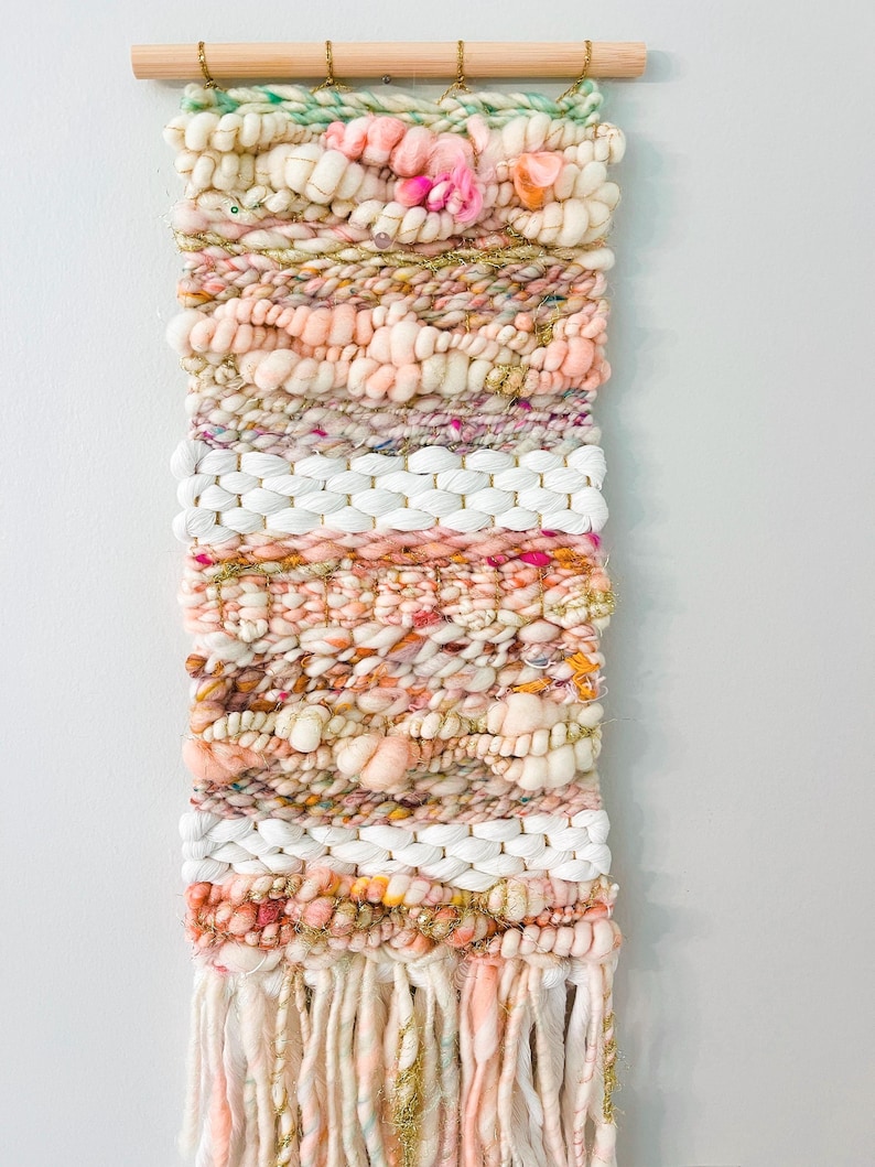 May include: A woven wall hanging with a pink, white, and gold color scheme. The hanging features a variety of textures, including fluffy yarn, chunky yarn, and thin yarn. The hanging is finished with a fringe of yarn.