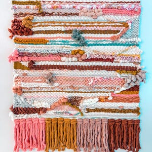 May include: A colorful woven wall hanging with a variety of textures and colors, including pink, brown, and white. The hanging has a fringe border and is made with a variety of yarns.