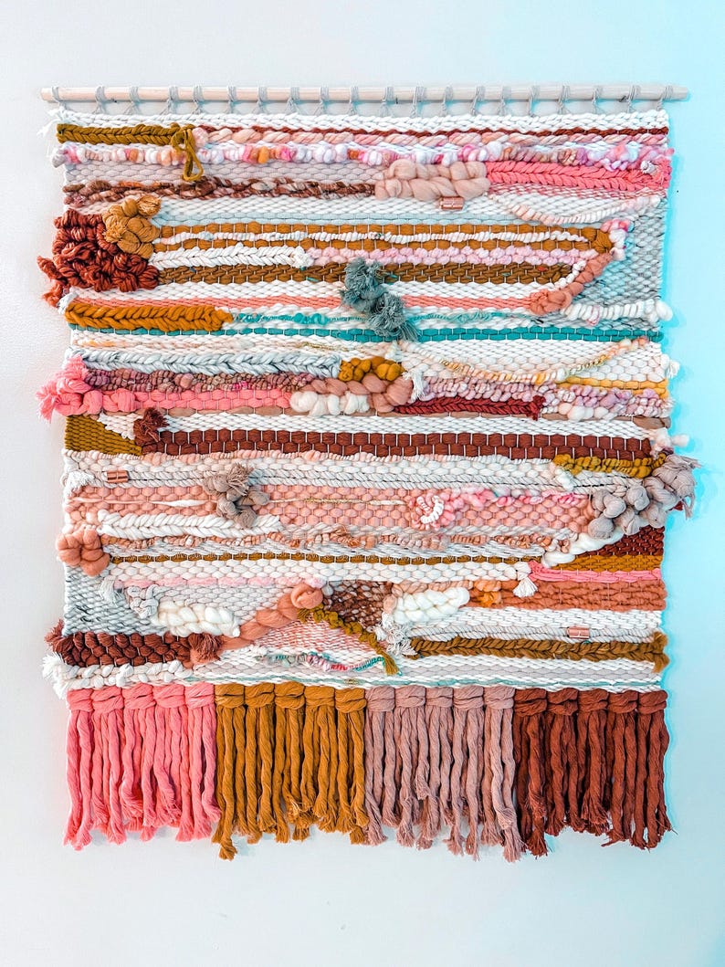 May include: A colorful woven wall hanging with a variety of textures and colors, including pink, brown, and white. The hanging has a fringe border and is made with a variety of yarns.