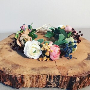 May include: A floral crown made with white, pink, and beige flowers, green leaves, and blue berries. The crown is resting on a wooden surface.