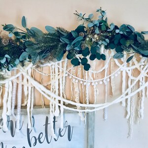 May include: A white yarn garland with white pom poms and lace trim hangs above a wooden sign with the text "Live Better". The garland is decorated with green eucalyptus leaves.