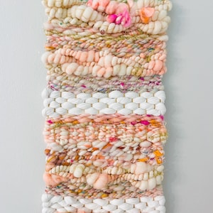 May include: A woven wall hanging with a pink, white, and gold color scheme. The hanging is made with a variety of textures and yarns, including fluffy yarn, sparkly yarn, and woven yarn.
