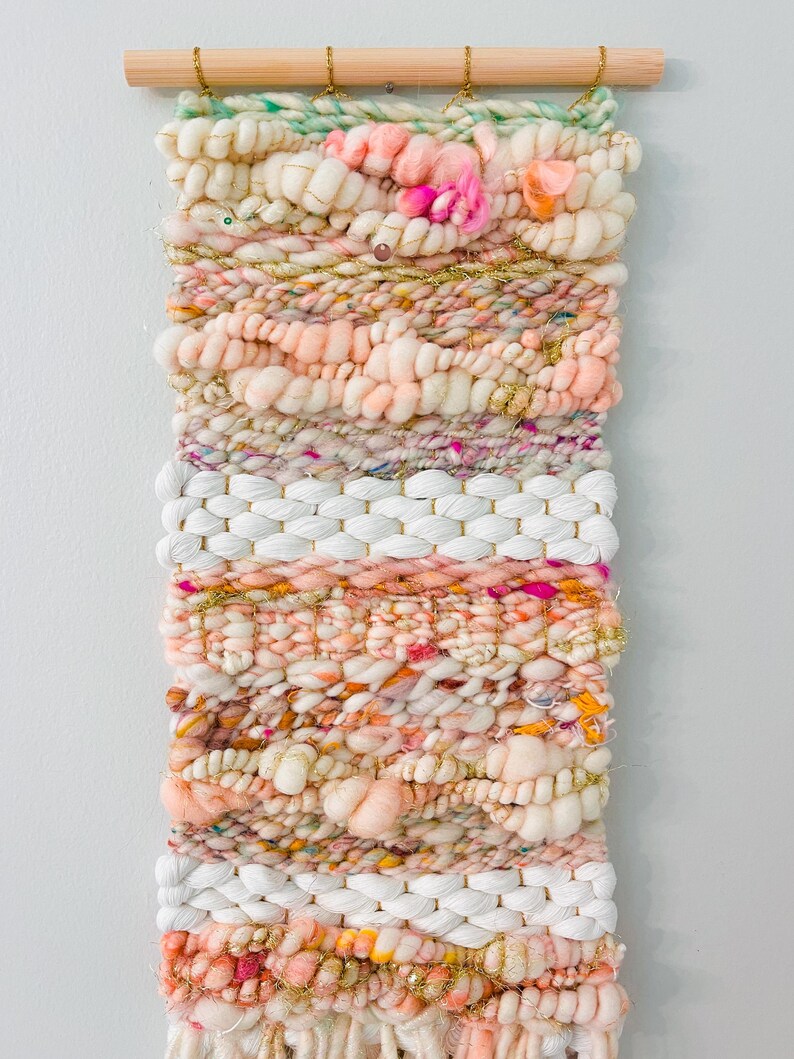 May include: A woven wall hanging with a pink, white, and gold color scheme. The hanging is made with a variety of textures and yarns, including fluffy yarn, sparkly yarn, and woven yarn.