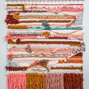 May include: A colorful woven wall hanging with a variety of textures and colors, including pink, orange, brown, and white. The hanging has a wooden dowel at the top and long fringe at the bottom.