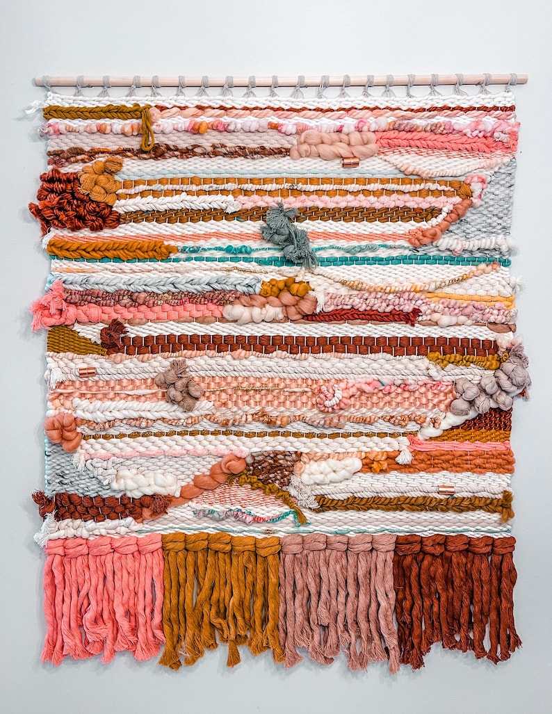 May include: A colorful woven wall hanging with a variety of textures and colors, including pink, orange, brown, and white. The hanging has a wooden dowel at the top and long fringe at the bottom.