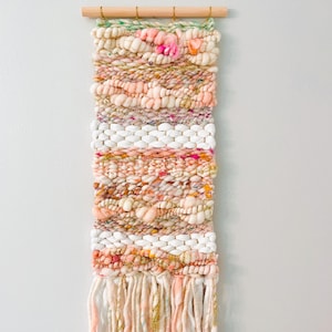 May include: A woven wall hanging with a pink, white, and gold color scheme. The hanging has a fringe at the bottom and is made with a variety of textures.
