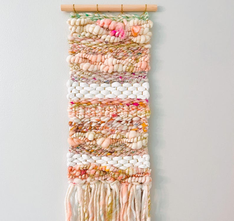 May include: A woven wall hanging with a pink, white, and gold color scheme. The hanging has a fringe at the bottom and is made with a variety of textures.