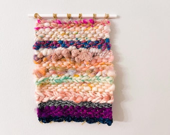 May include: A colorful woven wall hanging with pink, orange, green, blue, and purple yarn. The hanging is made with a variety of textures and patterns.