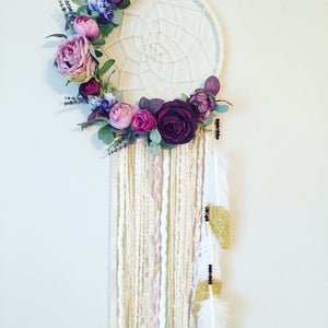 May include: A white dreamcatcher with a purple and pink floral wreath, white, gold, and pink yarn strands, and white feathers with gold glitter accents.
